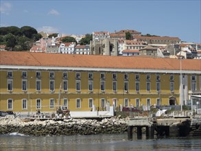 Coastal town with yellow buildings and a jetty by the water, surrounded by hills and houses,