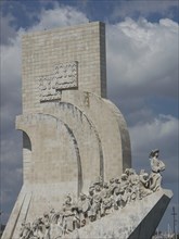 Large stone monument with sculptures of many figures under a partly cloudy sky, Lisbon, Portugal,