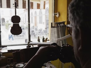 Silhouetted against a window, a luthier works on a stringed instrument in a workshop, with tools
