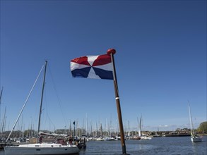 A water sports flag fluttering in the wind, boats and harbour facilities in the background under a