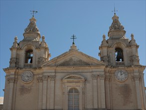 Front of a church with two heavily decorated bell towers and clocks, the town of mdina on the