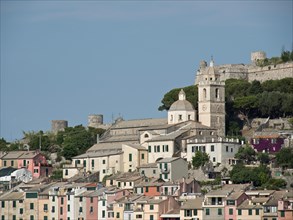 Church and fortress in a coastal town with colourful buildings in front of a wooded hill under a