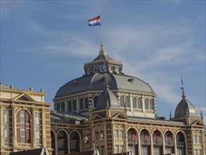 Historic building with Dutch flag in front of a blue sky, the beach of scheveningen with pier and