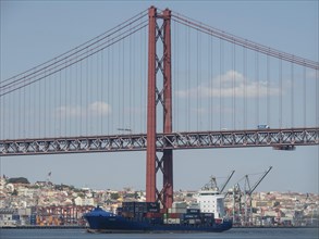 Red bridge over a river with a container freighter and city view in the background in Lisbon,