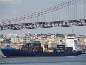 Large cargo ship loaded with containers under a bridge near the city on the waterfront, Lisbon,