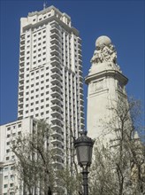 A statue in front of a skyscraper with trees and a lantern in the foreground under a blue sky,