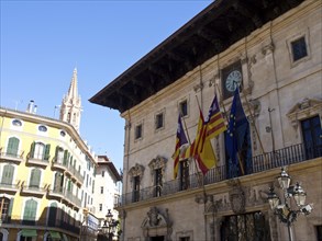 Historic building with various flags and a clock tower on a lively square under a clear sky, palma