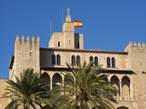 A historic building with a Spanish flag and palm trees in the foreground, under a clear blue sky,