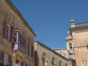 Row of historic buildings with balconies and shutters next to a bell tower, the town of mdina on