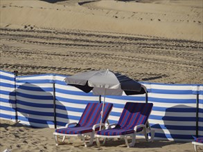 Blue and white striped deckchairs next to a parasol on the beach, bordered by blue and white