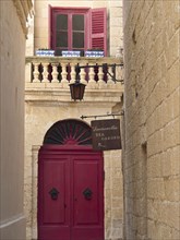 Narrow alley with a red door, a window and balconies above, the town of mdina on the island of