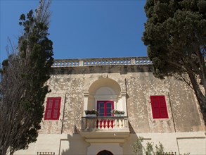 Buildings with red shutters and stone balconies, surrounded by trees, the town of mdina on the