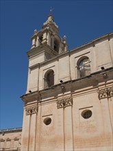 A tall church tower and sand-coloured buildings under a clear blue sky, the town of mdina on the