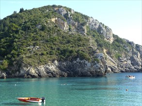 A picturesque coastal landscape with a green mountain and clear blue water in which a red boat