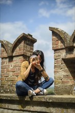 Thoughtful young woman sits on an old brick wall, casually dressed, enjoying a peaceful moment in a