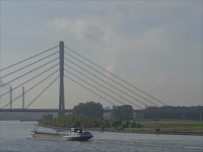 Ship sailing under a bridge in calm weather with clouds and trees along the shore, the Rhine near
