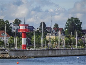 Red and white lighthouse and sailing boats along the coast against a background of trees and