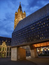 A modern building next to a historic church tower, both illuminated at night against a dark sky,