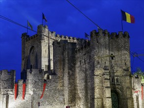 Imposing illuminated castle with several waving flags in front of a deep blue night sky, blue hour