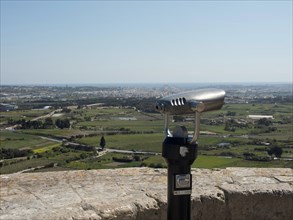 A pair of binoculars at a viewpoint overlooking a vast landscape and a distant city, the town of