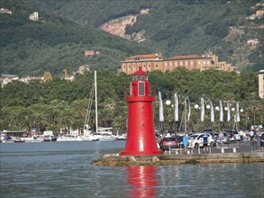 A red lighthouse at the harbour with boats, surrounded by trees and mountains in the background,