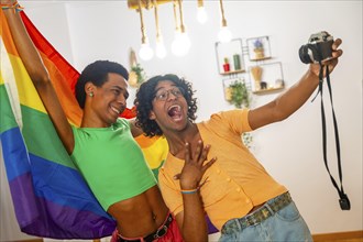 Gay latin young couple taking a selfie with a camera while raising rainbow lgbt flag at home