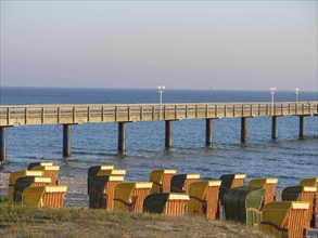 Beach chairs on the beach in front of a long pier leading into the sea, in the warm colours of the