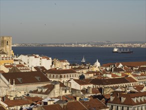 Expansive city view with red roofs, behind it the sea and a ship under a clear sky, Lisbon,