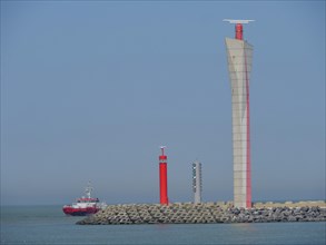A lighthouse and a red ship in front of a coast with concrete blocks in a clear blue sky,