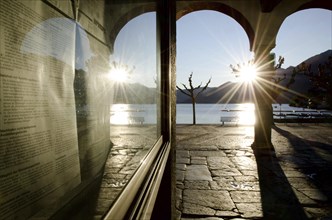 Alley with Arches on the Waterfront to Lake Maggiore with Mountain and Sunbeam and Reflection on a