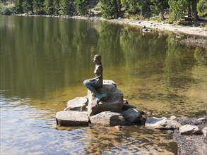 Statue of a seated figure on rocks on the shore of a quiet lake, small forest lake in the black