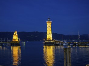 Illuminated lighthouse and lion statue at night, whose lights are reflected in the water of the