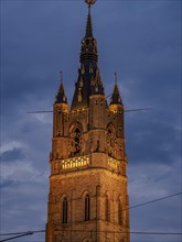 High church tower with clock and coloured lights at dusk, blue hour in a medieval town with