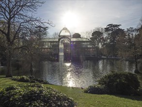 Sunlight hits a greenhouse by a lake, surrounded by trees and reflected in the water, Madrid,