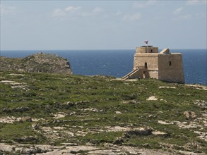 Historic watchtower on a green, rocky coastal area under a blue sky with a flag, green field with
