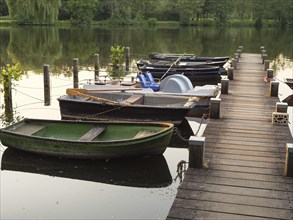 A jetty leads to several boats floating on a calm lake, green trees and bushes on a small lake with