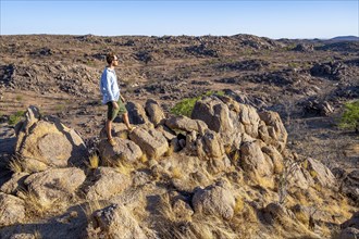 Young man on rocks, barren landscape with rocky hills and acacia trees, African savannah in the