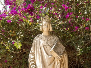 Stone statue of a king with book and cross surrounded by blooming pink flowers, Tunis in Africa