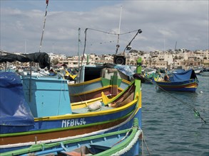 Several fishing boats in the harbour of a town, traditionally painted under a blue sky, many