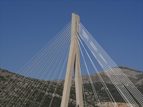 Modern suspension bridge with ropes in front of a clear blue sky and mountains, the old town of