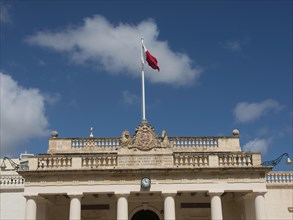 A historic building with a flag on the roof, surrounded by columns, under a blue sky, Valetta,
