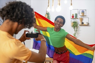Gay man taking photos while couple posing waving LGBT rainbow flag at home