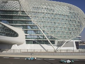Two racing cars drive alongside a modern building with a lattice structure on the race track in