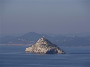A rocky island with a lighthouse, surrounded by a calm sea and hills, the old town of Dubrovnik