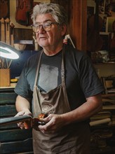 A craftsman wearing glasses and an apron works on a violin in a workshop, illuminated by a desk
