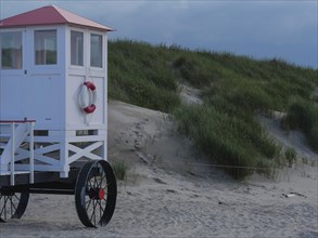 A white rescue tower with a red roof stands on a sandy beach in front of green dunes, Baltrum