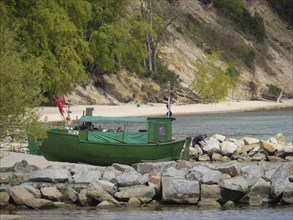 A green boat lies on a rocky shore, in the background dense forest and sandy beach, spring on the