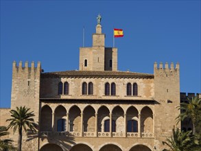 Historic building with a Spanish flag on the tower, surrounded by palm trees and battlements,