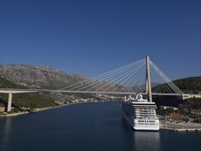 Suspension bridge over a river with a large cruise ship, surrounded by mountains and blue sky, the