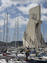 Large monument next to a lively harbour with many sailing boats under a cloudy sky, Lisbon,